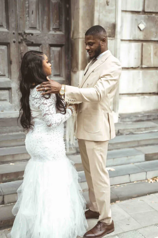 An elegant bride in a lace mermaid gown and groom in a beige suit share a tender glance on stone steps outside a classic building.