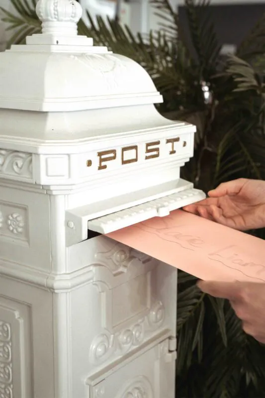 A close-up shot of hands posting a pink card into a vintage white post box decorated with lettering.