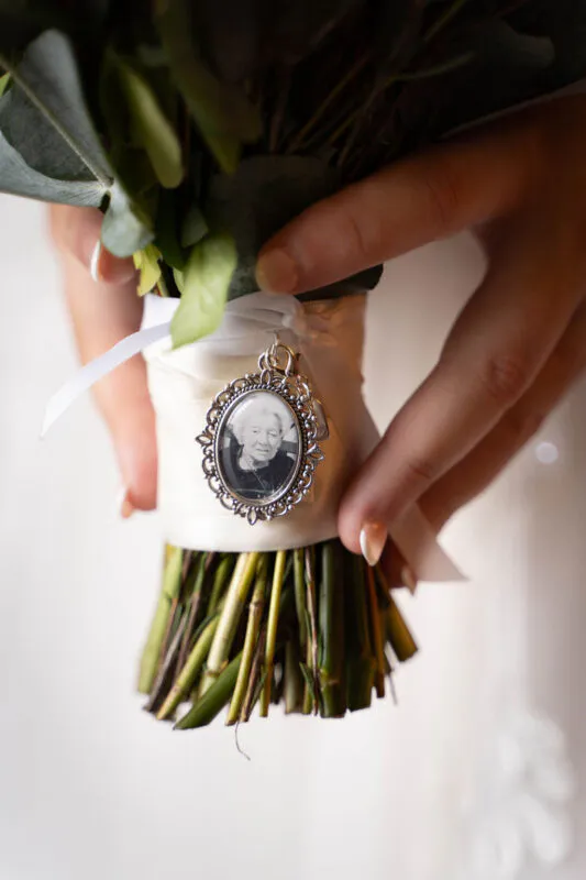 Close-up of a bride holding a bouquet wrapped in white ribbon, featuring a vintage silver locket containing a sentimental family photograph pinned to the stems.