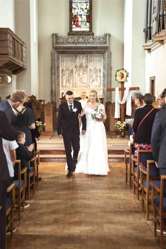 Newlywed bride and groom walk hand in hand down the aisle of a historic church during their recessional, smiling as guests applaud from wooden pews.