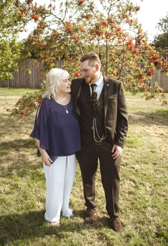 An elderly woman smiling and looking up at a groom wearing a brown tweed suit as they stand together outdoors near a tree with red berries.