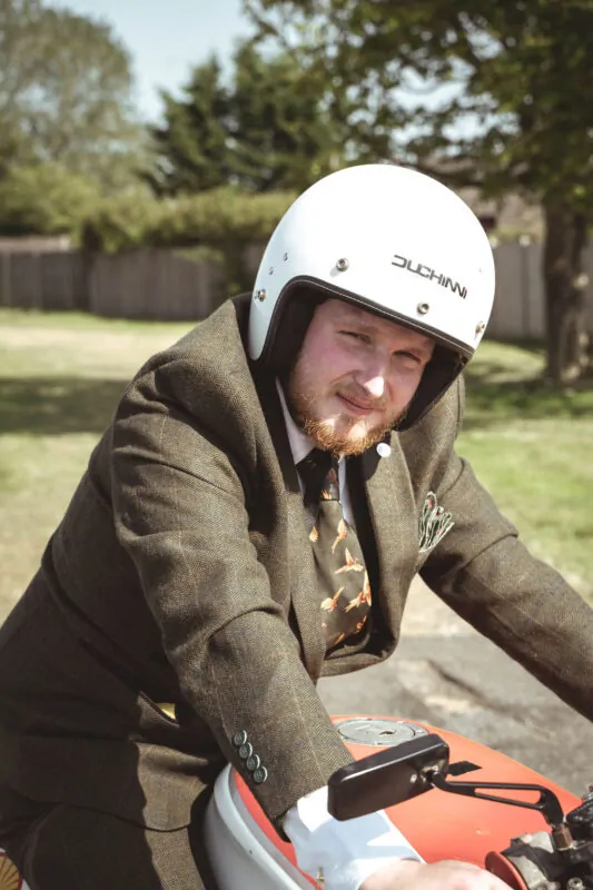 A groom wearing a tweed suit and a white motorcycle helmet sits on a motorbike outdoors.