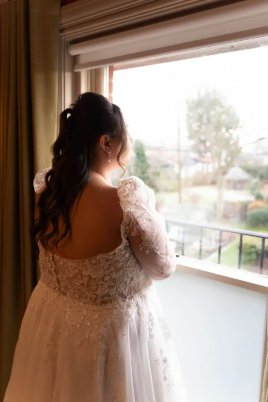 A bride stands by a window in a white lace gown with long sheer sleeves, looking outside during morning preparations.