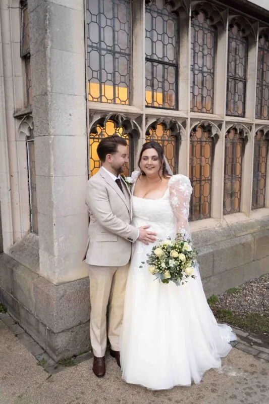 A newlywed couple poses together outside a historic stone building with leaded glass windows, with the groom looking lovingly at the bride who holds a cream floral bouquet.