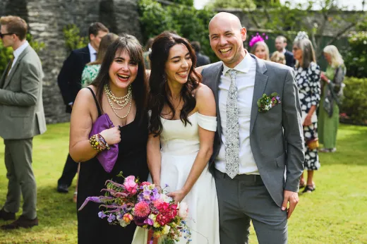 A joyful bride in an off-the-shoulder gown holding a vibrant wildflower bouquet laughs alongside her groom and a guest during a sunny outdoor reception.