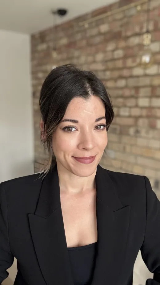 A close-up portrait of a woman with dark hair in a low bun, wearing a black blazer against an exposed brick wall.