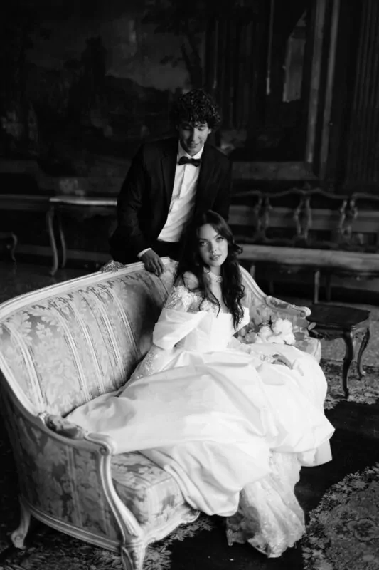 A newlywed couple poses together in an opulent stately home, with the bride seated on an antique floral sofa in an off-the-shoulder white gown while the groom stands behind her in a classic black tuxedo.