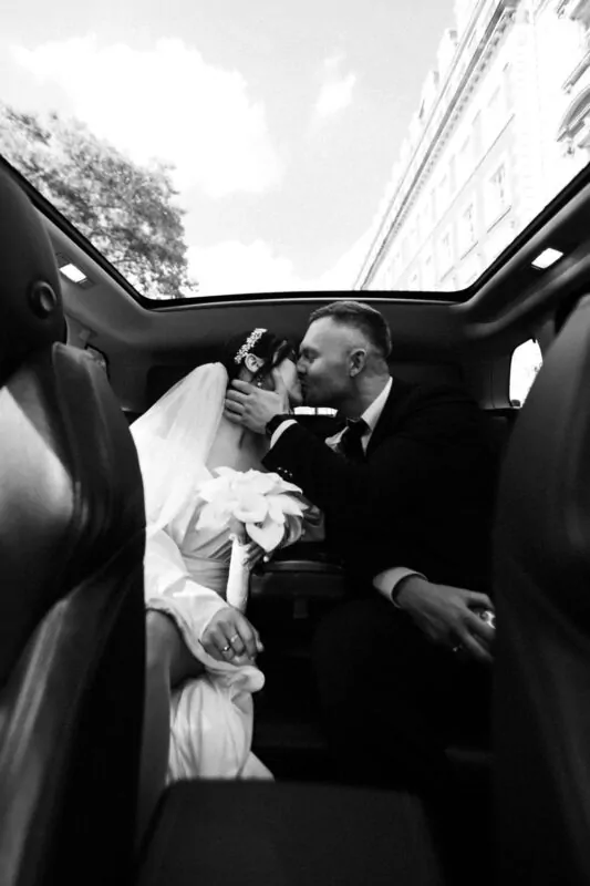A newlywed couple share a tender kiss in the back of a luxury car with a panoramic glass roof as they travel through the city on their wedding day.