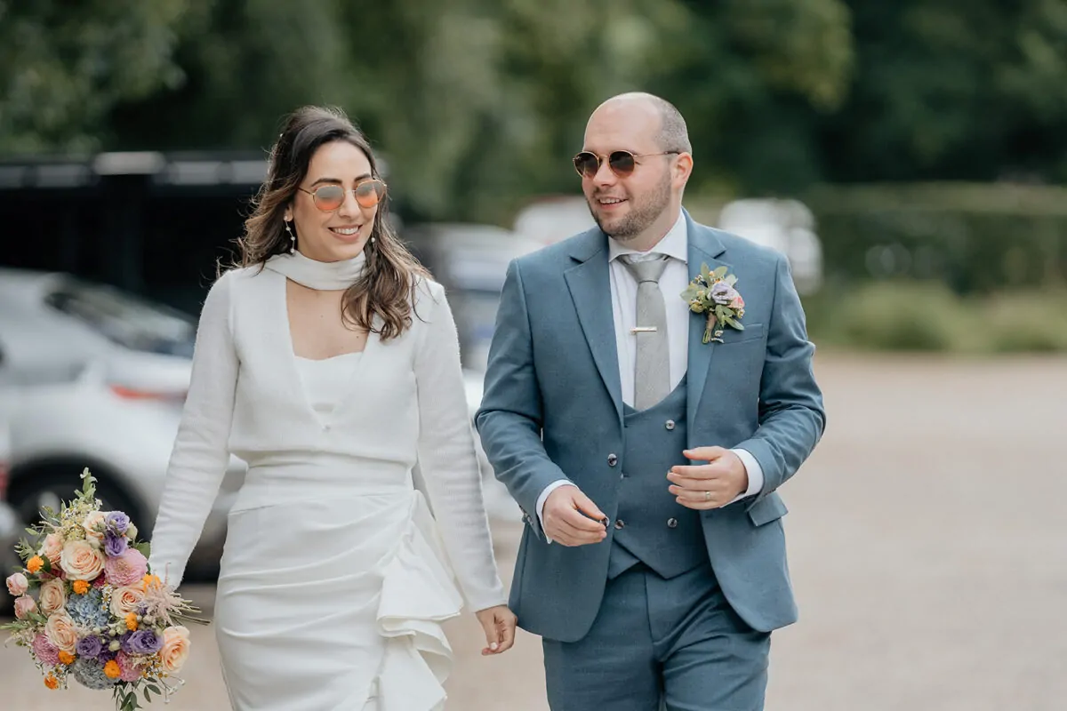 A stylish newlywed couple wearing sunglasses walk happily outdoors, with the bride carrying a vibrant mixed flower bouquet and wearing a white dress.