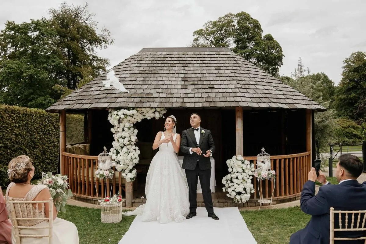A smiling bride and groom release a white dove outside a wooden gazebo decorated with white floral arrangements during their outdoor wedding ceremony.
