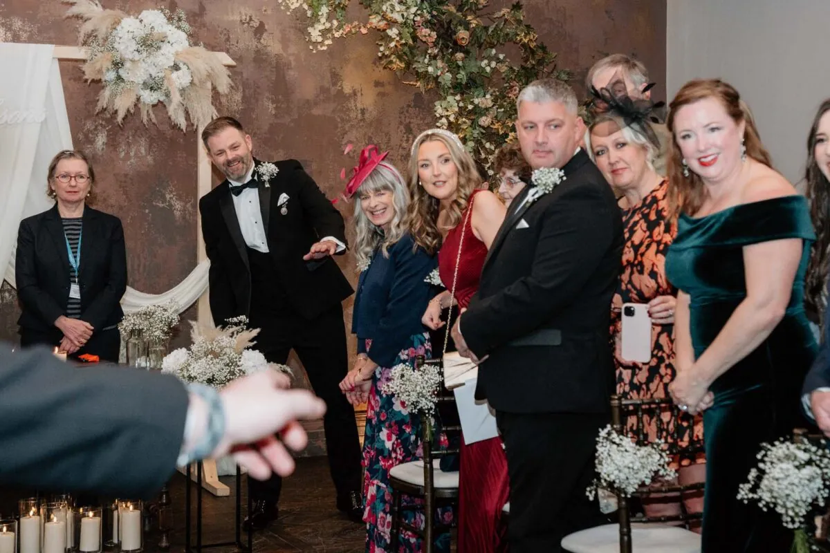 A group of smiling guests and wedding party members watch joyfully during an indoor wedding ceremony lit by direct flash.