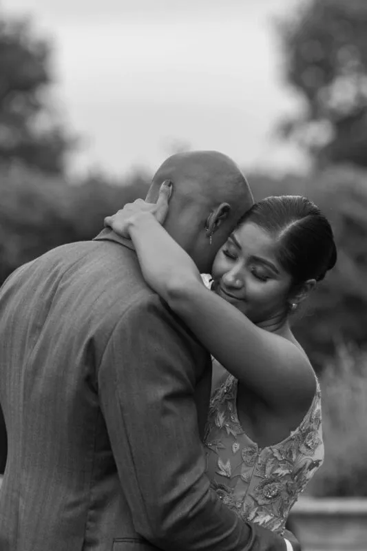 A black and white portrait captures a tender embrace between a newlywed couple, with the bride resting her head against the groom's shoulder.