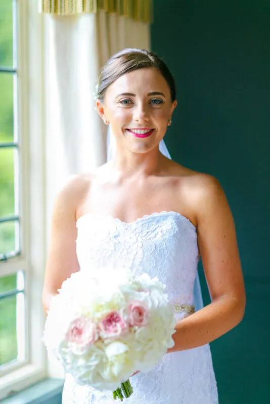A smiling bride wearing a lace strapless wedding gown and holding a round pink and white rose bouquet stands near a bright window.