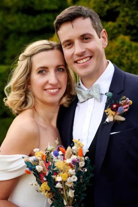 A smiling bride and groom pose closely together outdoors, showcasing their colourful dried flower bouquet and matching boutonniere against a green foliage backdrop.