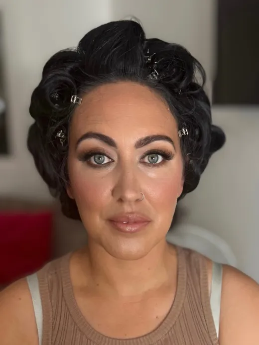 A close-up portrait of a bride getting ready with hair rollers and pins in her dark hair while wearing soft glam makeup.
