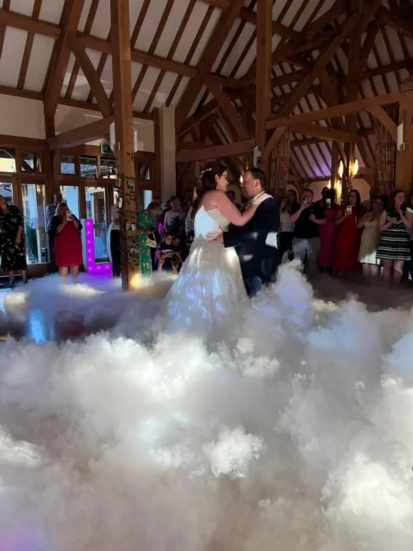 A newlywed couple shares their first dance on a dance floor surrounded by low-lying fog inside a rustic oak-framed barn with guests watching.