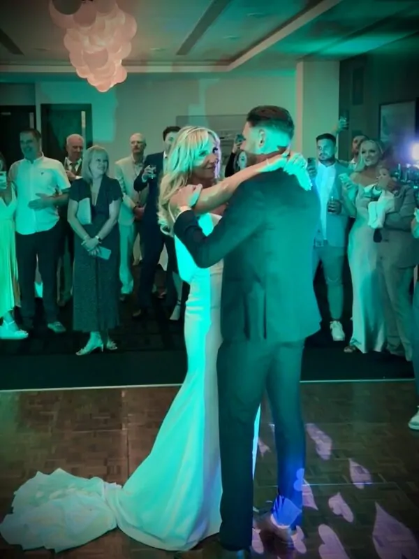 The newly married couple share their first dance together on a wooden dance floor surrounded by cheering guests under atmospheric green and blue lighting.