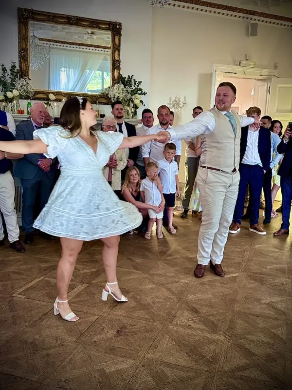 Newlyweds share their first dance with arms outstretched on a parquet floor inside a grand manor house ballroom while guests watch from the sidelines.