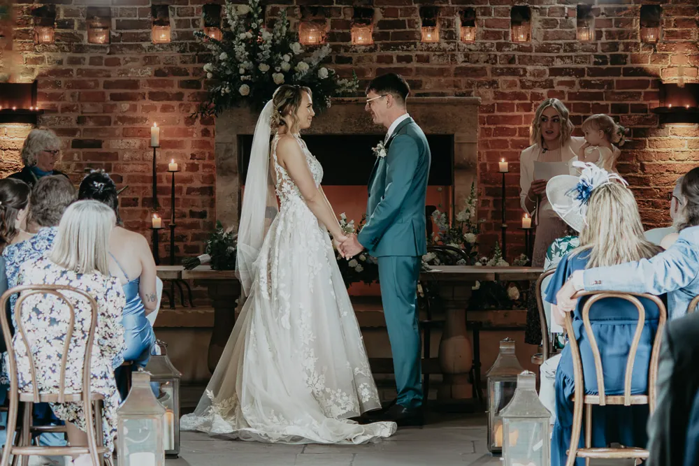 A bride in a lace appliqued gown and groom in a green suit exchange vows during an indoor ceremony framed by exposed brick and candles.