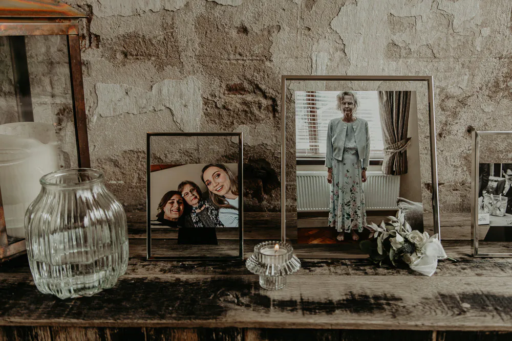 A rustic memory table features framed family photographs displayed alongside a glass vase, a lit votive candle, and a small floral arrangement against an exposed brick wall.