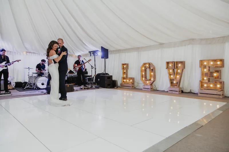 Newlyweds share their romantic first dance on a white dance floor inside a draped marquee with a live band playing in the background.