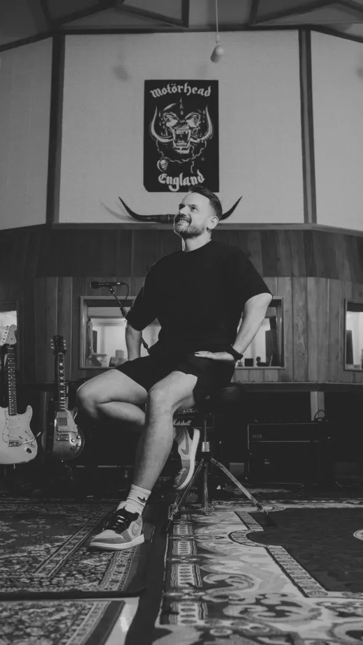 A groom in a black t-shirt and shorts smiles while sitting on a stool inside a music recording studio surrounded by guitars and a motorhead banner.