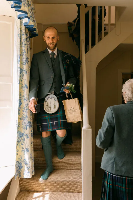 A smiling groom wearing a traditional kilt and tweed jacket walks down a carpeted staircase carrying a paper bag.