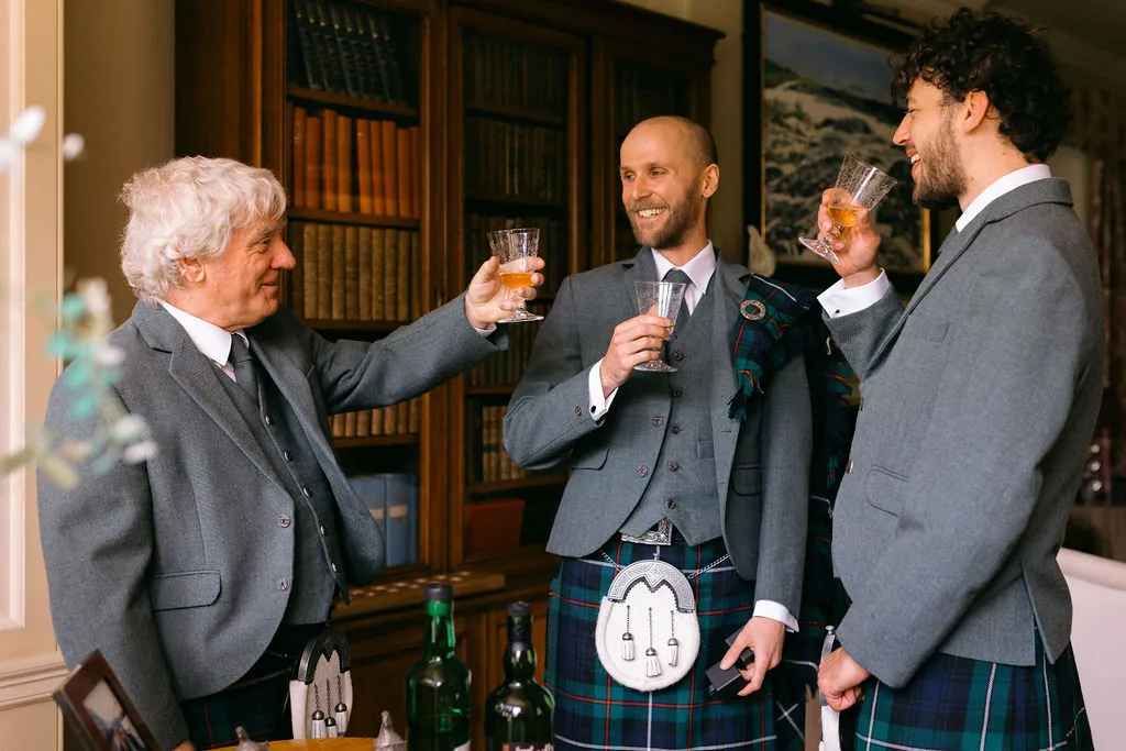 Three men in traditional highland dress and matching grey tweed jackets raise their glasses in a toast inside a wood-panelled library.