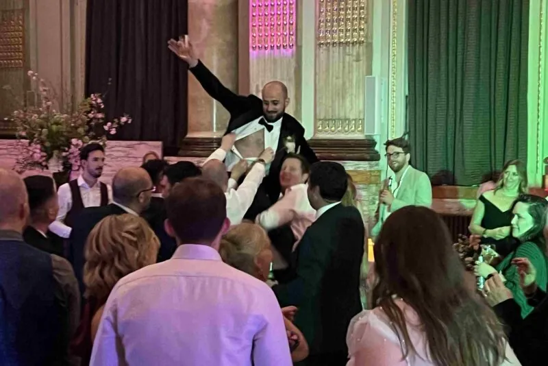 A groom is lifted into the air by guests on a packed dance floor during an energetic evening wedding reception in a grand ballroom.