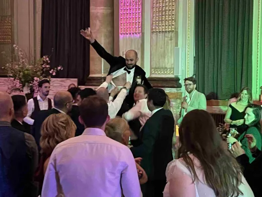 A groom is lifted into the air by guests on a packed dance floor during an energetic evening wedding reception in a grand ballroom.