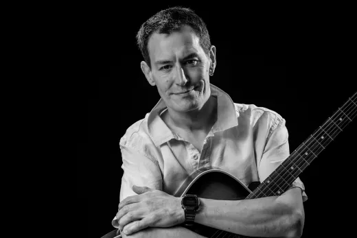 A musician poses with an acoustic guitar in a dramatic black and white studio portrait.