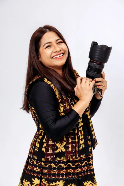 A smiling woman with long brown hair wearing a patterned traditional vest holds a professional camera against a plain white background.