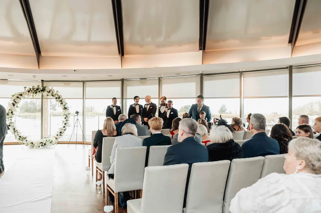 A wide angle interior shot of a wedding ceremony inside a modern venue featuring a circular floral arch and seated guests watching from the light-filled room.