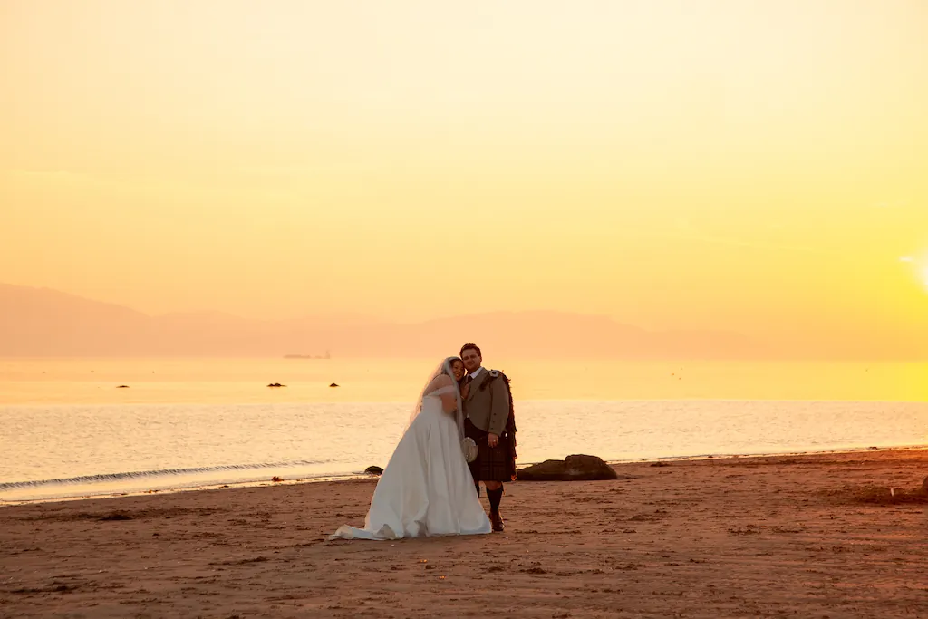 A newlywed couple poses together on a sandy beach during a breathtaking golden sunset over the calm sea.