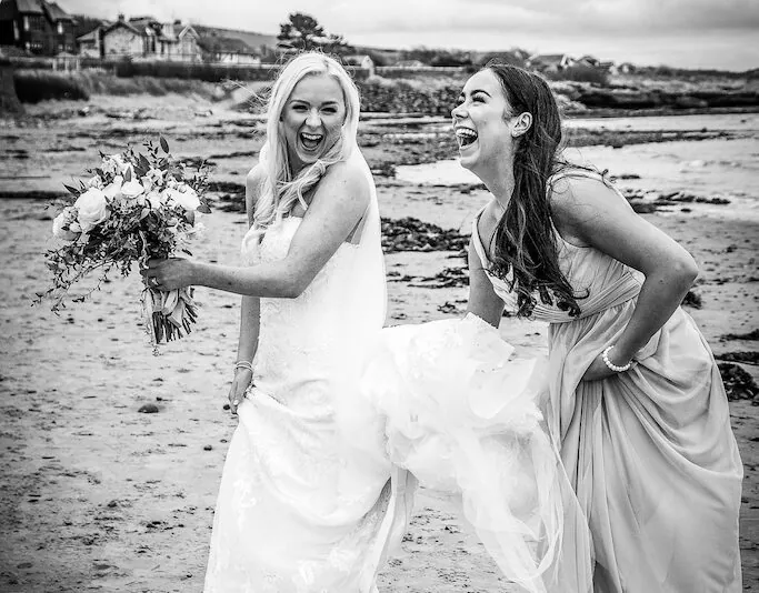 A joyful bride in a white gown and sliders laughs hysterically on a pebbled beach alongside her bridesmaid, holding a hand-tied bouquet.