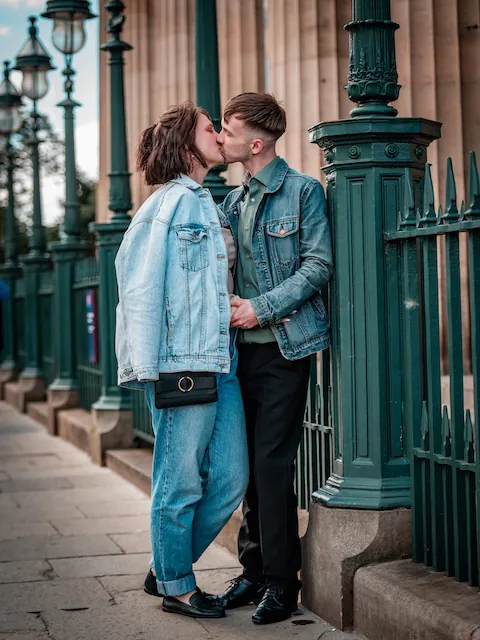 A newlywed couple shares a tender kiss on a city street next to ornate green iron railings and vintage streetlamps.