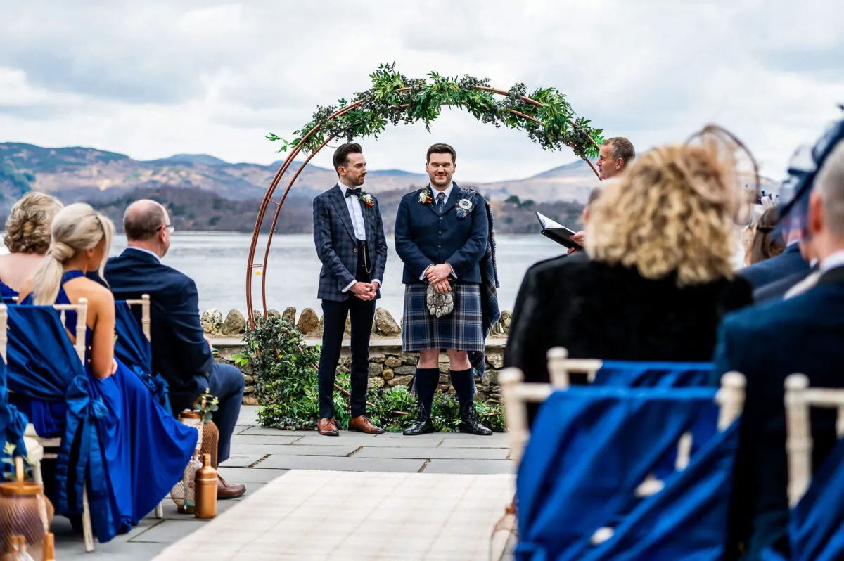 Two grooms stand beneath a circular floral arch during an outdoor lakeside ceremony with mountain views as guests watch from seated rows.