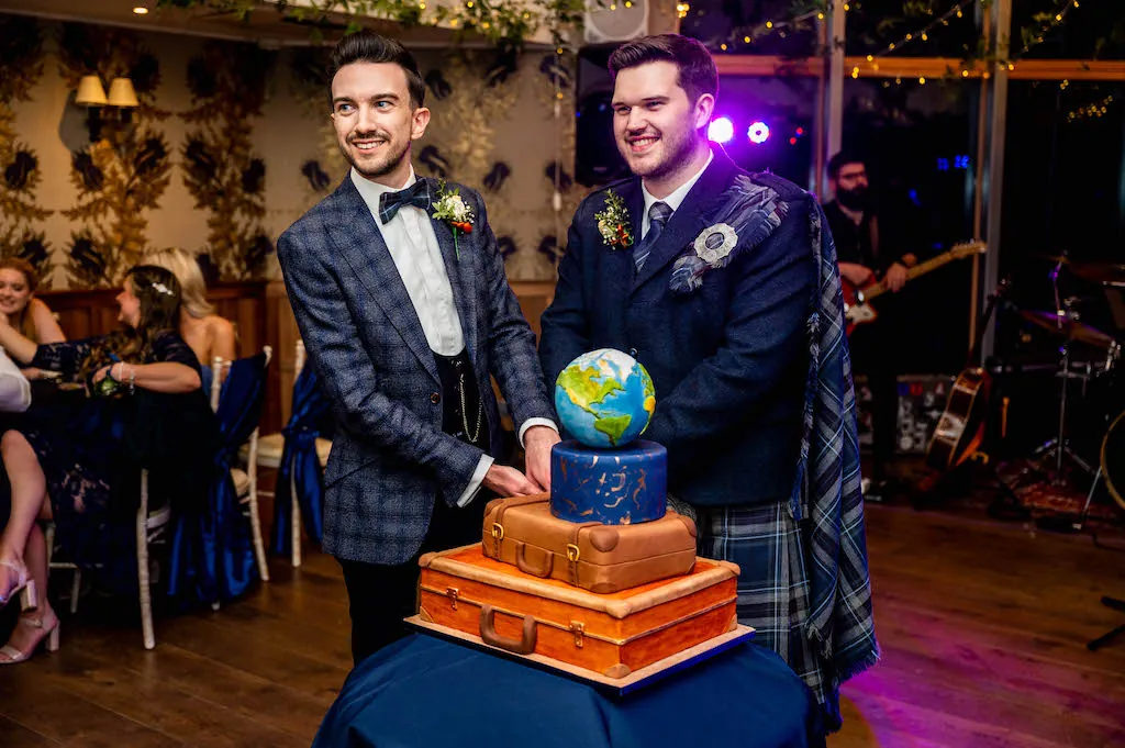 Two grooms proudly cut their travel-themed wedding cake shaped like stacked vintage suitcases topped with a globe, standing together at their evening reception.