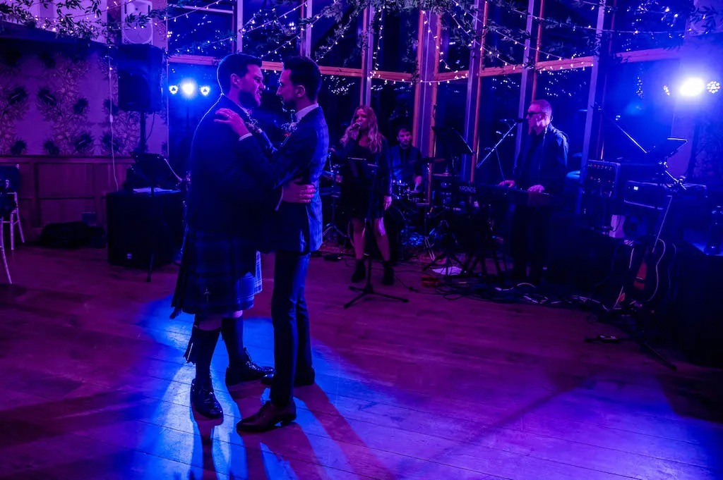 Two grooms share their first dance on a wooden dance floor illuminated by vibrant blue stage lighting while a live band performs in the background.