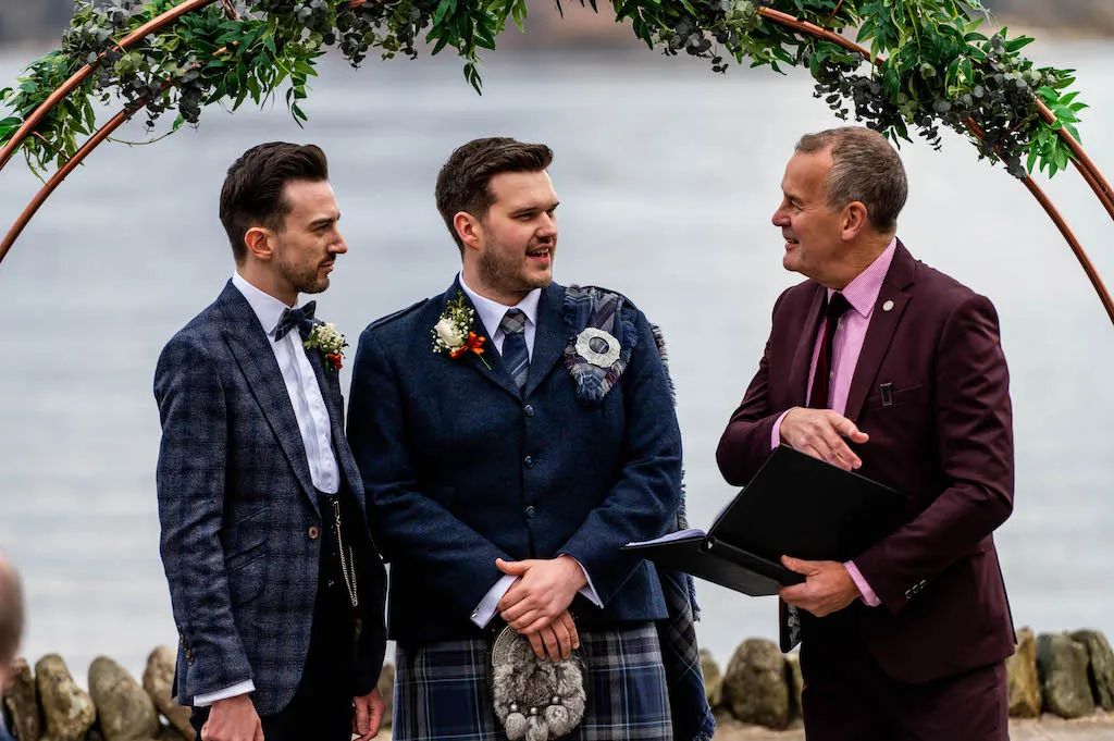 Two grooms in patterned suits and a kilt stand beneath a copper arch adorned with greenery during an outdoor lakeside ceremony, conversing with the celebrant.