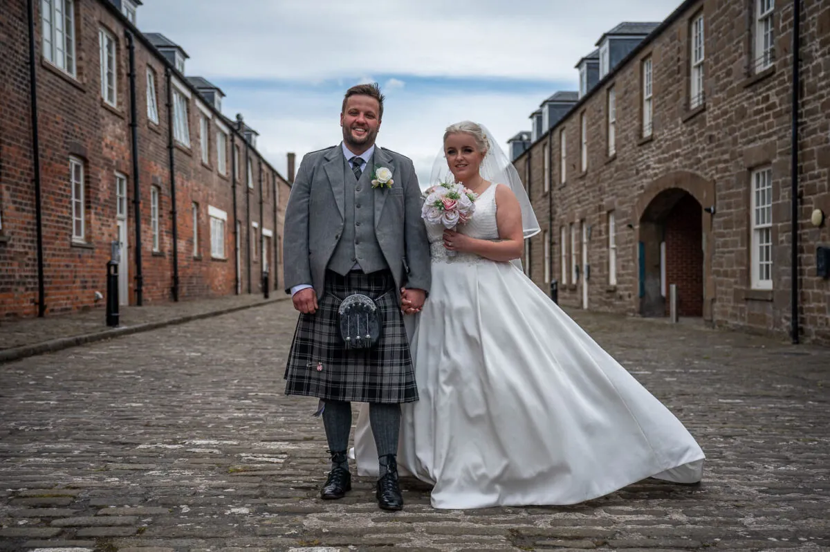 A newlywed couple posing hand in hand along a historic cobbled street lined with traditional stone buildings, the groom wearing a traditional kilt and tweed jacket while the bride wears an elegant A-line wedding dress and veil.