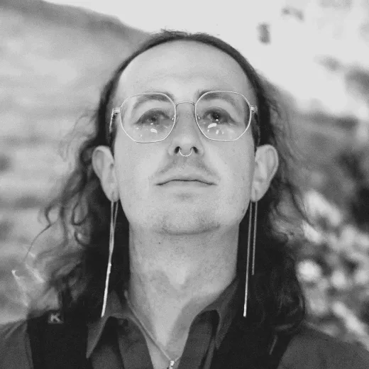 A striking black and white portrait of a long-haired person wearing wire-rimmed glasses, long drop earrings, and a septum ring.