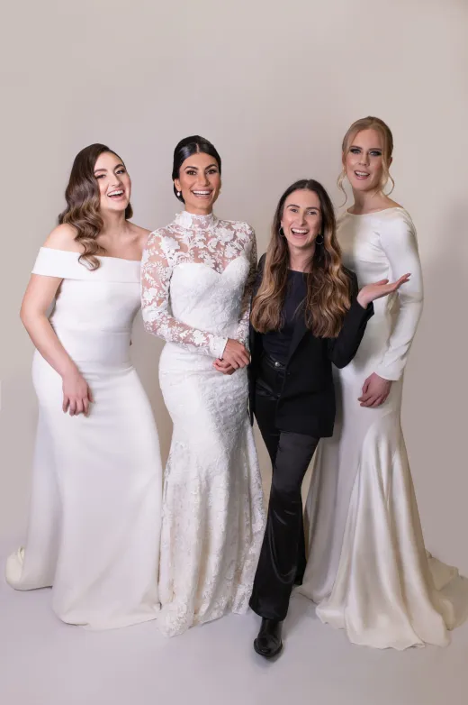 A smiling bride in a lace high-neck gown stands proudly alongside two bridesmaids in white dresses and a laughing stylist in a black suit.