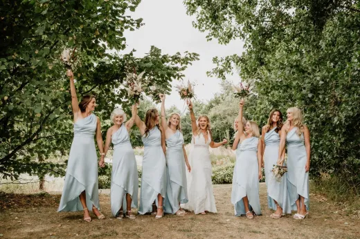 The smiling bride and her bridesmaids wearing matching satin dusty blue one-shoulder dresses lift their bouquets in celebration outdoors amongst lush green trees.
