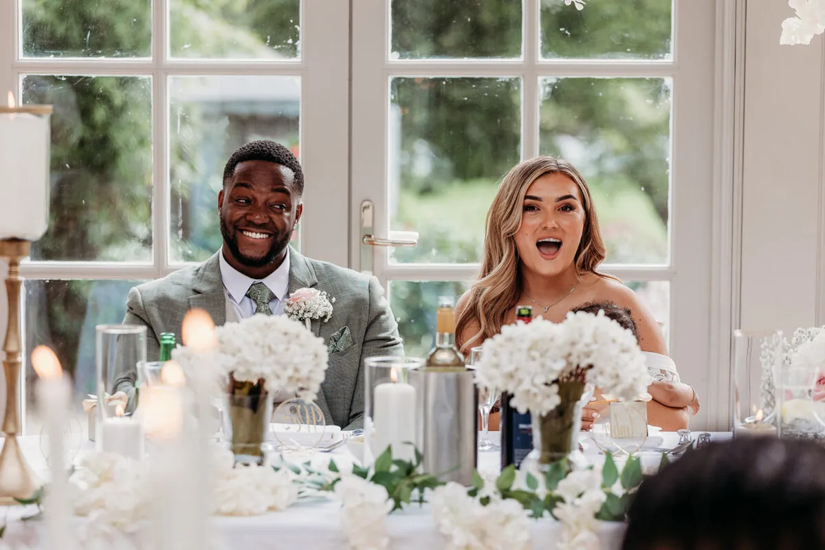 An interracial newlywed couple laugh and react joyfully during wedding speeches at a top table decorated with white floral arrangements and glowing candles.