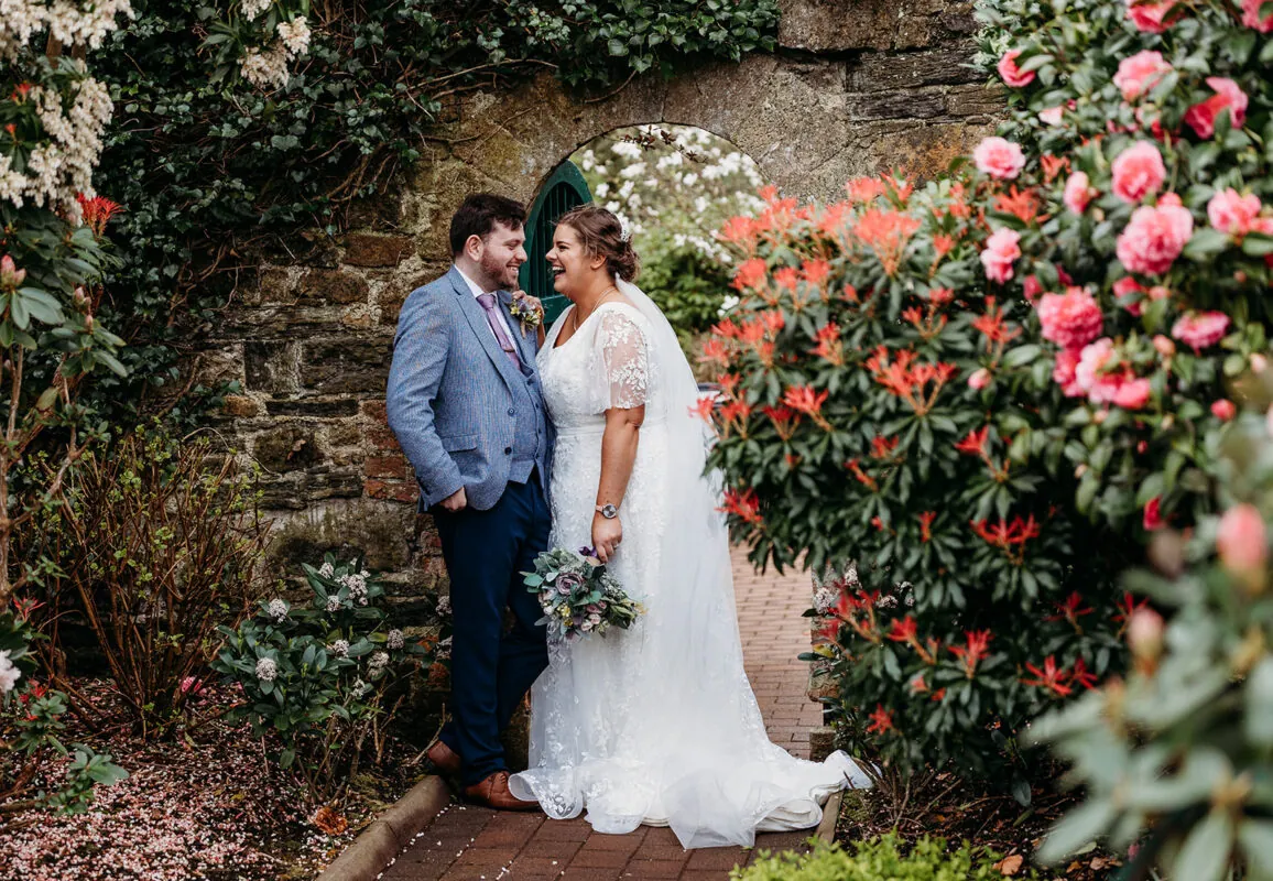A happy newlywed couple laughing together framed by a historic stone archway and lush garden foliage during their outdoor wedding portrait session.