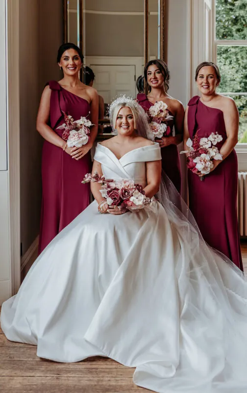 A smiling bride seated in a voluminous satin off-the-shoulder gown holds a floral bouquet while three bridesmaids in matching one-shoulder burgundy dresses stand behind her.