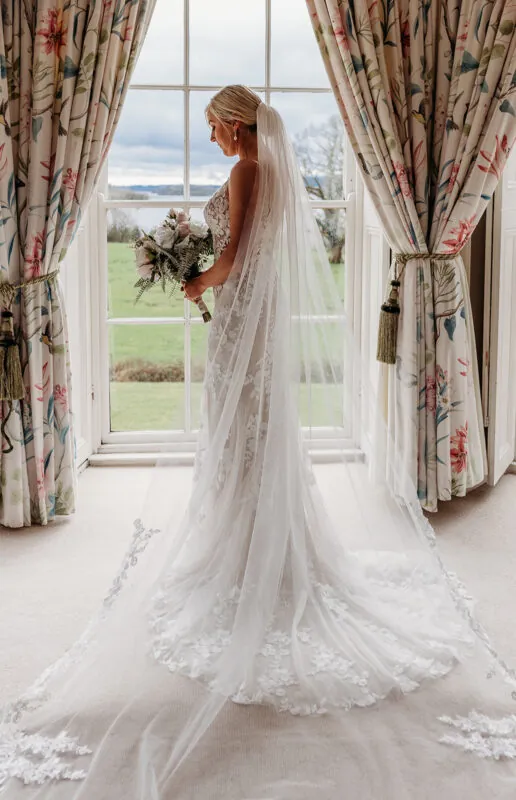 A stunning bride stands by a large window holding a bouquet, wearing a lace gown and a flowing veil that pools on the carpet.