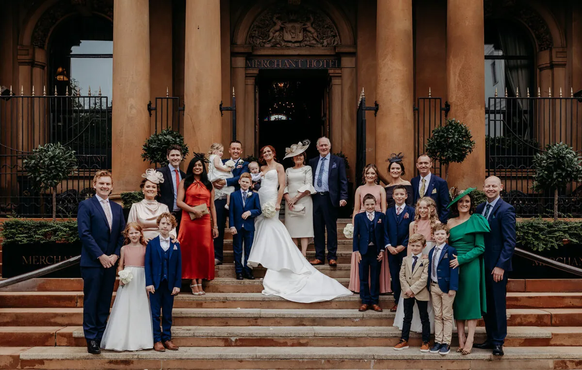 A large wedding party and family members pose together on the grand stone steps outside the entrance of the Merchant Hotel.