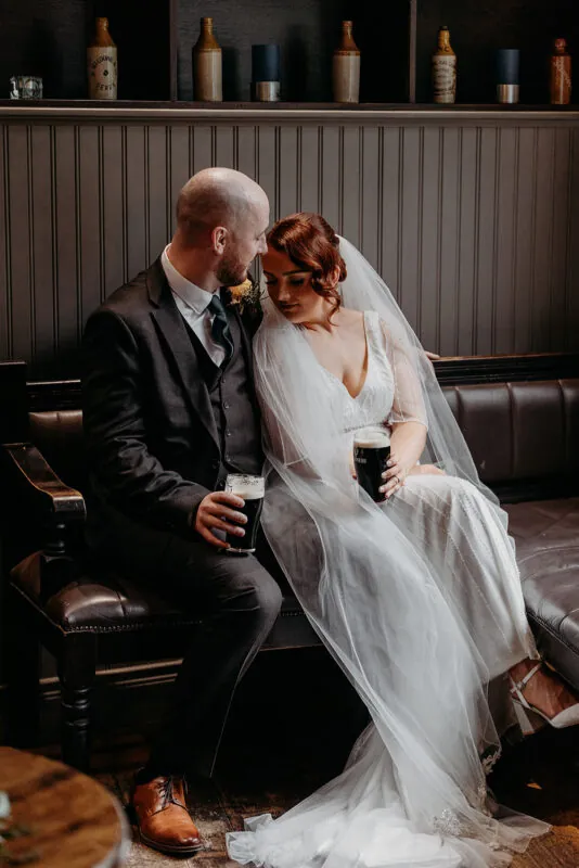 A newlywed bride and groom sit closely together on a leather banquette inside a cosy pub, each holding a pint of Guinness while enjoying a quiet moment.
